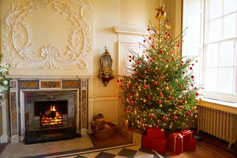 A gorgeously dressed Christmas tree surrounded by red-wrapped present stands on a black and white chequered floor. The fire is crackling in the grate in an exquisite marble fireplace beneath a swirling rococo wall plaster design. The log basket is full and a small gilded rococo mirror reflects the dancing lights on the Christmas tree. This is the entrance hall at Paxton House ready for Christmas.