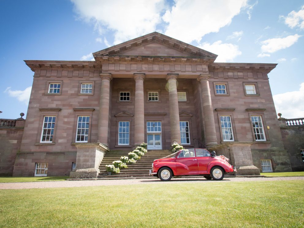 A tall elegant Palladian facade features a pillared portico and symmetrical Georgian windows set into a warm read sandstone house. The steps are decorated with groups of white flowers and in front stands a bright red beetle car all ready for a wedding