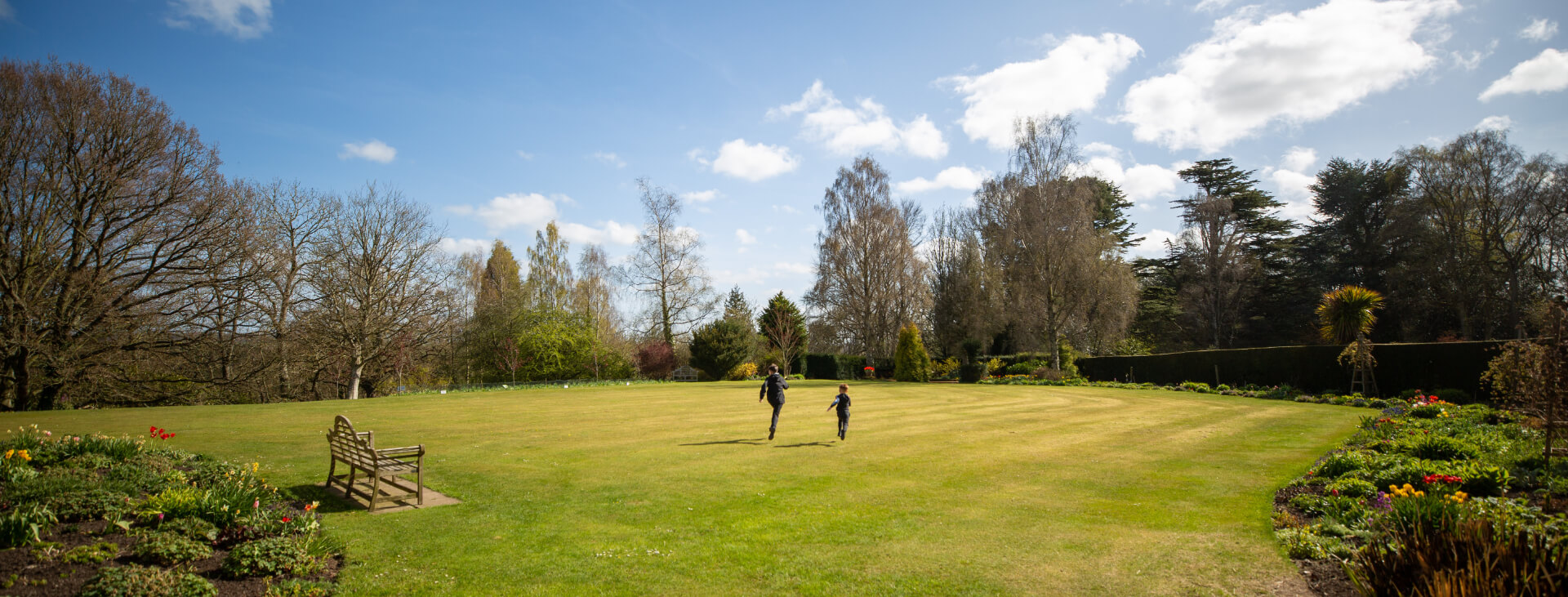 Two boys are running across a wide expanse of lawn leading to a range of trees