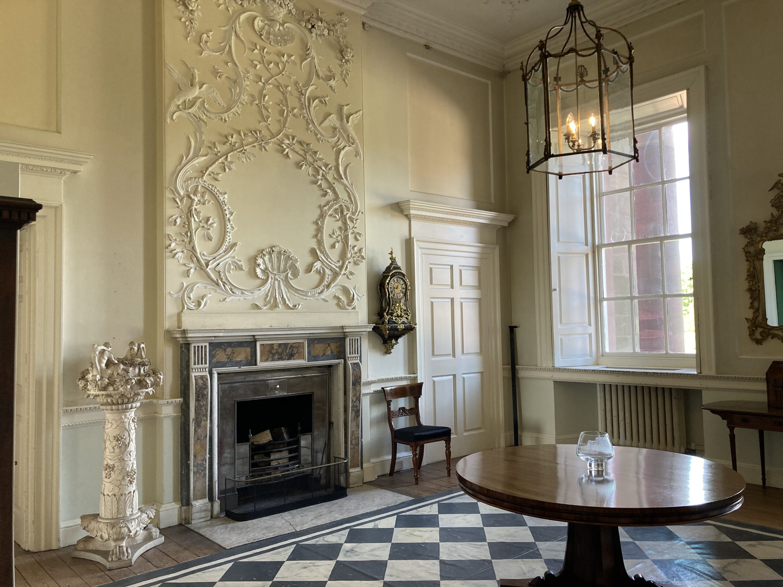 A Georgian room with a black and white painted mat on the floor is bathed in sunshine. The fireplace wall is covered in decorative Rococo plasterwork and a collar mahogany tiles stands in the foreground beneath a large metal and glass lantern.