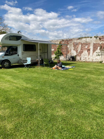 A dark haired girl sits reading on a rug on a rich sward of green lawn. She is dressed in a navy top, soaking up the sun just beyond the shade cast by a simple awning supported by a pole and attached to the side of a white camper van. Behind her are pillows and she is clearly relaxed and at ease. Under the awning are camping chairs and a table. The camper van stands in a walled garden, the walls behind are old and scarred, with mellow brickwork randomly splashed with old lime wash and ivy.