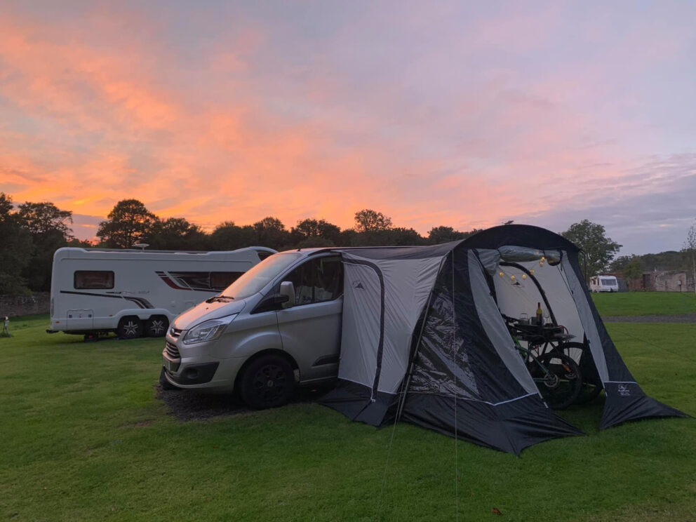 A camper van with its awning spread sits in the gloaming with a glowing orange sunset sky behind it