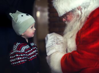 A small smiling boy in a fair isle sweater and a bobble hat stands in front of Father Christmas who is leaning forward to offer the boy a wrapped Christmas present. He has a red velvet coat and hat edged with white fur and a long white beard. 