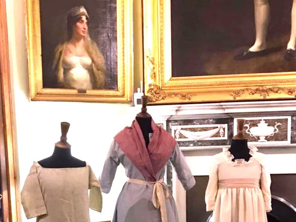 Three manikins in costume stand in a period room before a marble fireplace. Above the fireplace hadn't two portraits. We can just the see legs of a gentleman in britches and black leather shoes. We can see the full portrait of a lady in Regency dress with dark hair, looking away from us.