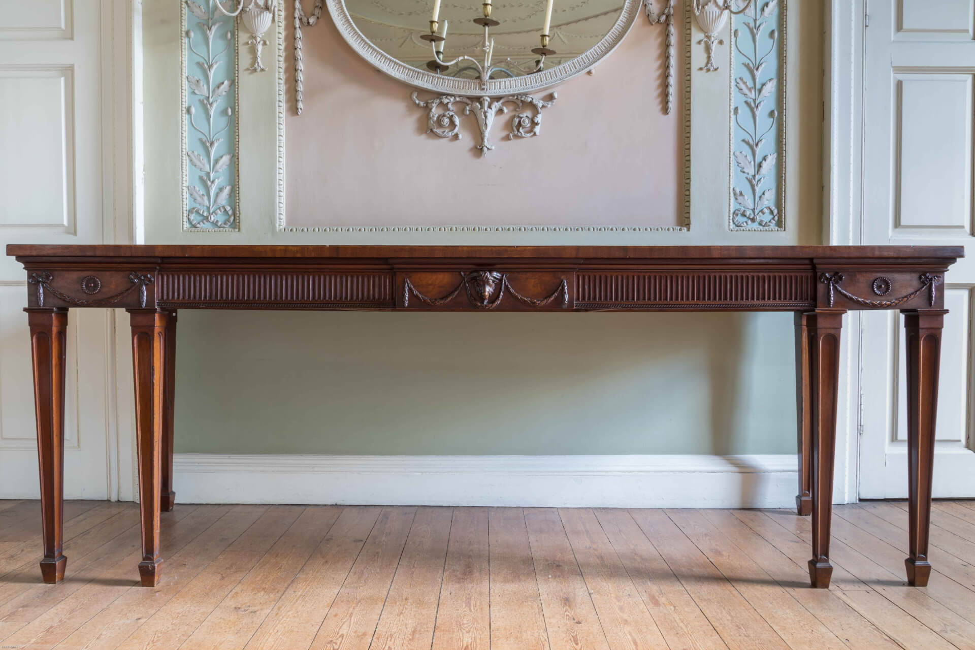 A period interior in green and pink with an ornate rococo oval mirror finished with a two candle sconce above a narrow sideboard table. The table, by Thomas Chippendale is decorated with ormolu masks and ribbons. flanking it are two urn cellarettes with delicate curving bronze handles.