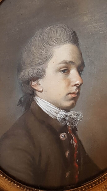 a boy in a powdered wig looks sideways in this profile portrait. He is wearing a white stock tied around his neck and a brown wool coat. This is Patrick Home of Billie