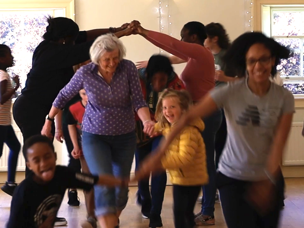 A group of people of diverse heritage, adults and children are dancing traditional Scottish reels. Two of the dancers make an arch with their hands and the others a squeezing through the space hand in hand. They are laughing and having fun.