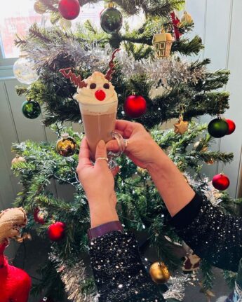 Someone is holding up a Christmas milk shake with a reindeer face made from cherries. chocolate chips and flake in the froth on the top. Behind is a decorated Christmas tree.