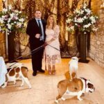 A wedding couple stand in the Victorian boathouse, decorated with fishing nets lights ad plinths supporting large bouquets of flowers. They are holding tightly to the leads of two bulldogs which are nosing around the floor.