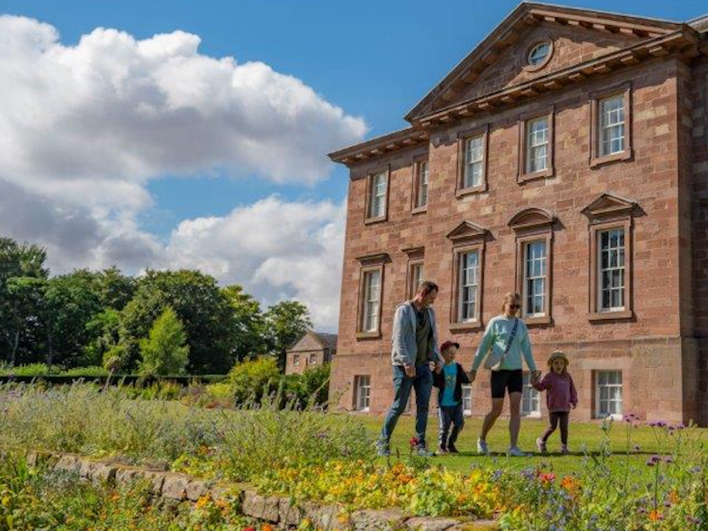 A mother, father and two young children are walking cheerfully along the lawn in front of a red sandstone classical house. To their left is a colourful border planted in yellows and purples.