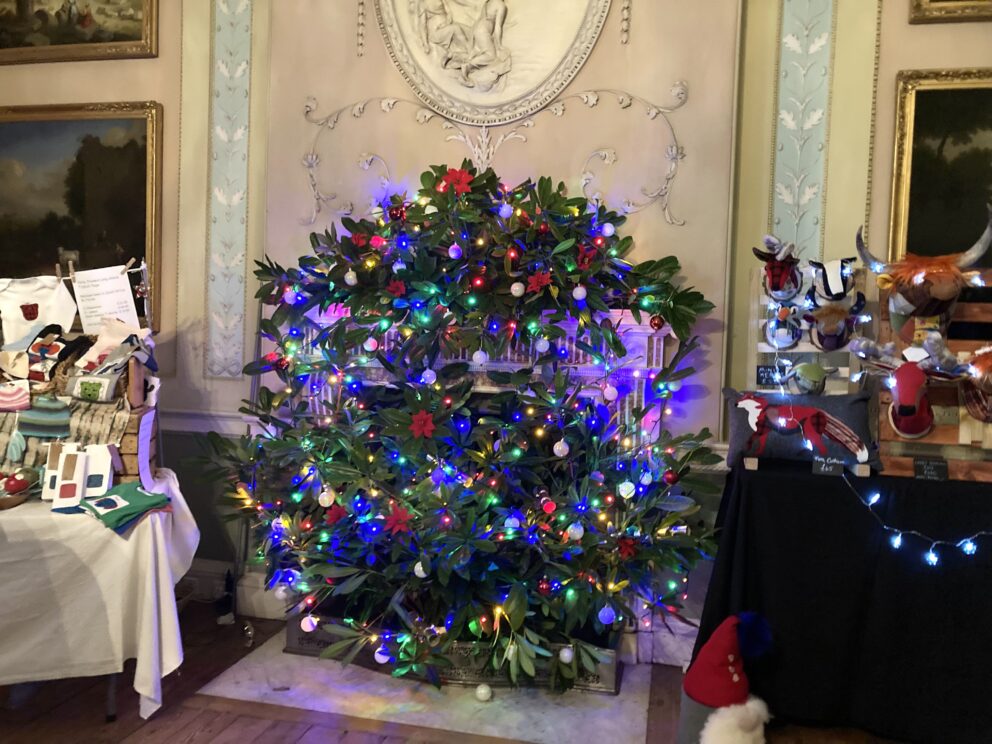 A huge Christmas wreath fills a fireplace It is covered in blue Christmas lights and silvery bows. The fireplace is delicately decorated with Rococo plasterwork. Just out of frame are a Christmas tree and a pile of Christmas parcels.