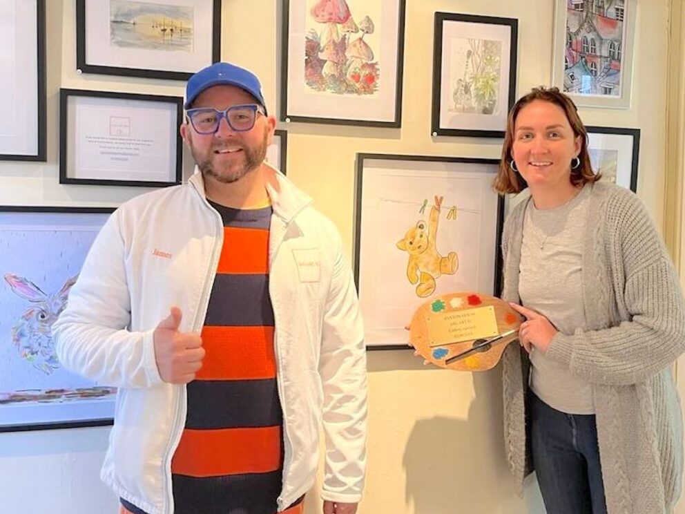 A bearded man in a brilliantly striped shirt and a blue baseball cap gives a cheery thumbs up next to a smiling woman with bobbed hair holding a colourful artists pallette. They stand in front of a wall covering in watercolour images of nature, landscape and animals including Paxton Ted.