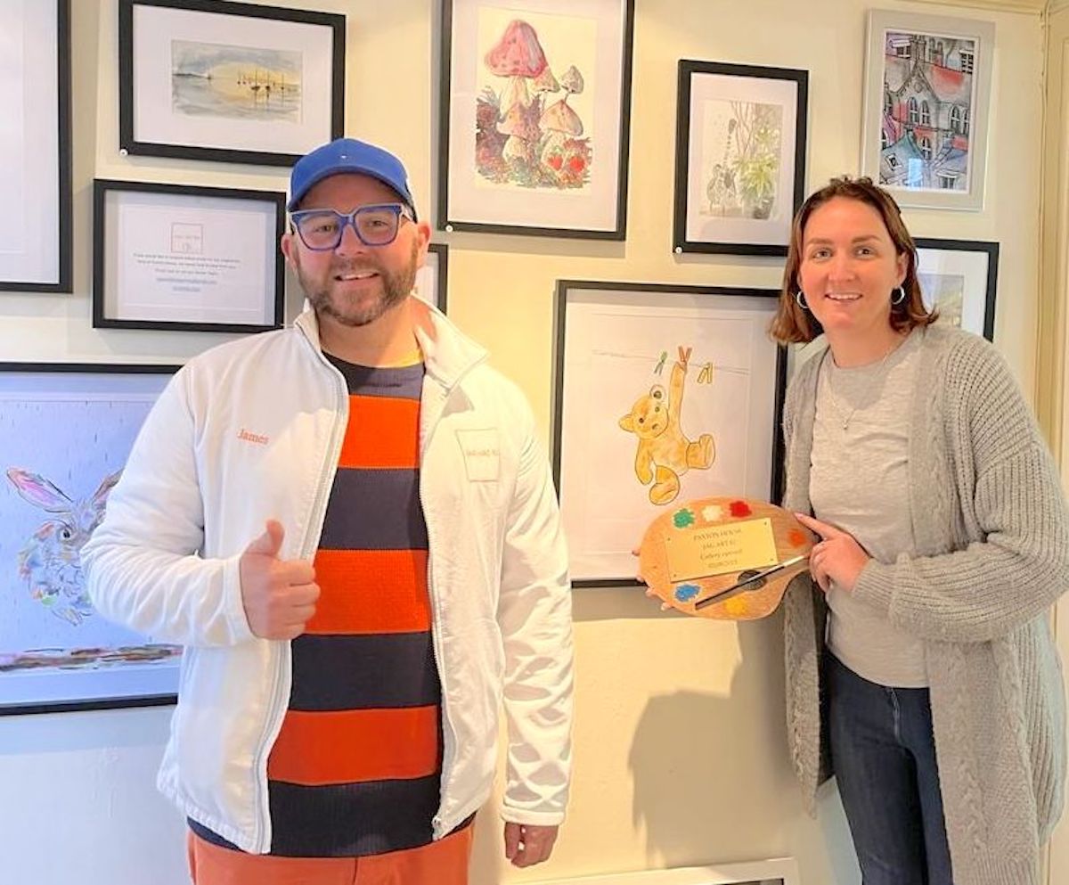A bearded man in a brilliantly striped shirt and a blue baseball cap gives a cheery thumbs up next to a smiling woman with bobbed hair holding a colourful artists pallette. They stand in front of a wall covering in watercolour images of nature, landscape and animals including Paxton Ted.