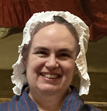 The head and shoulders of smiling woman dressed in a cotton mob cap with her hair pulled back out of sight looks into the camera