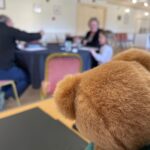 The back of furry teddy's bear's head is in the foreground. The bear is apparently watching three people - child of about 8, mother and male artist playing with paints and brushes at a small table.