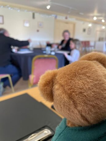 The back of furry teddy's bear's head is in the foreground. The bear is apparently watching three people - child of about 8, mother and male artist playing with paints and brushes at a small table.