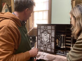 A skilled restored stands. looking at an inlaid ivory and rosewood cabinet. The door has small decorated ivory panels around a large coat of arms. The interior is s set of drawers around a small ivory figurine of the god Hercules. It stand on a surface covered with a brownish red cloth and the restored holds a dusting brush in his hand.