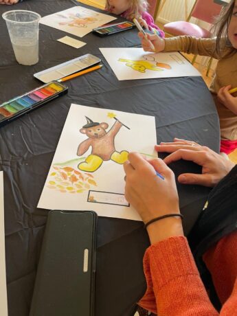  A watercolour painting of a teddy bear dressed as a magician takes shape on a piece of white paper set against a black background. The artist's hands, pencil and orange jersey sleeves are visible as they draw and hold down the edge of the paper. A small tin of watercolour paints sits towards the top of the frame.