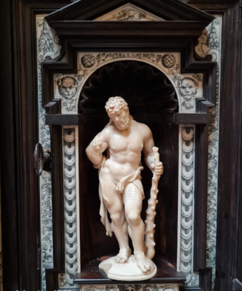 An ivory statuette of a man in a loincloth holding a knobbly staff is Hercules, Demi-god of Greek myth. He is looking down to the right and is heavily muscled. He stands in a dark wood niche decorated with inlayed and inscribed ivory in a renaissance style. 