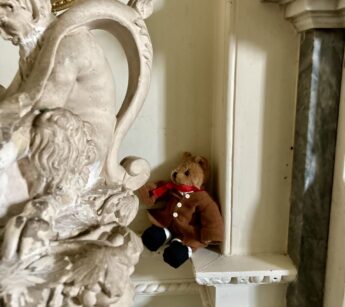 A small smartly dressed teddy bear dies in the corner of the fireplace in the Entrance Hall at Paxton House. He is sitting on the dado rail. In the foreground id the ornate handle of a plaster vase featuring a satyr with furry legs. The corner of an oval picture frame is just visible on the wall behind.