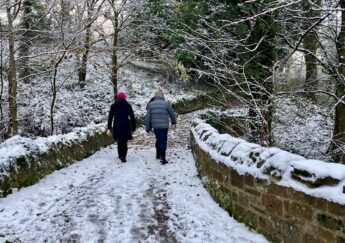 A couple walk together on a path lightly sprinkled with snow.  They are walking between two low stone walls through woods along the Linn Burn at Paxton House. Tehe black branches of the trees are edged with frost. The walkers are well wrapped up in hats and anoraks against the cold and walking briskly.