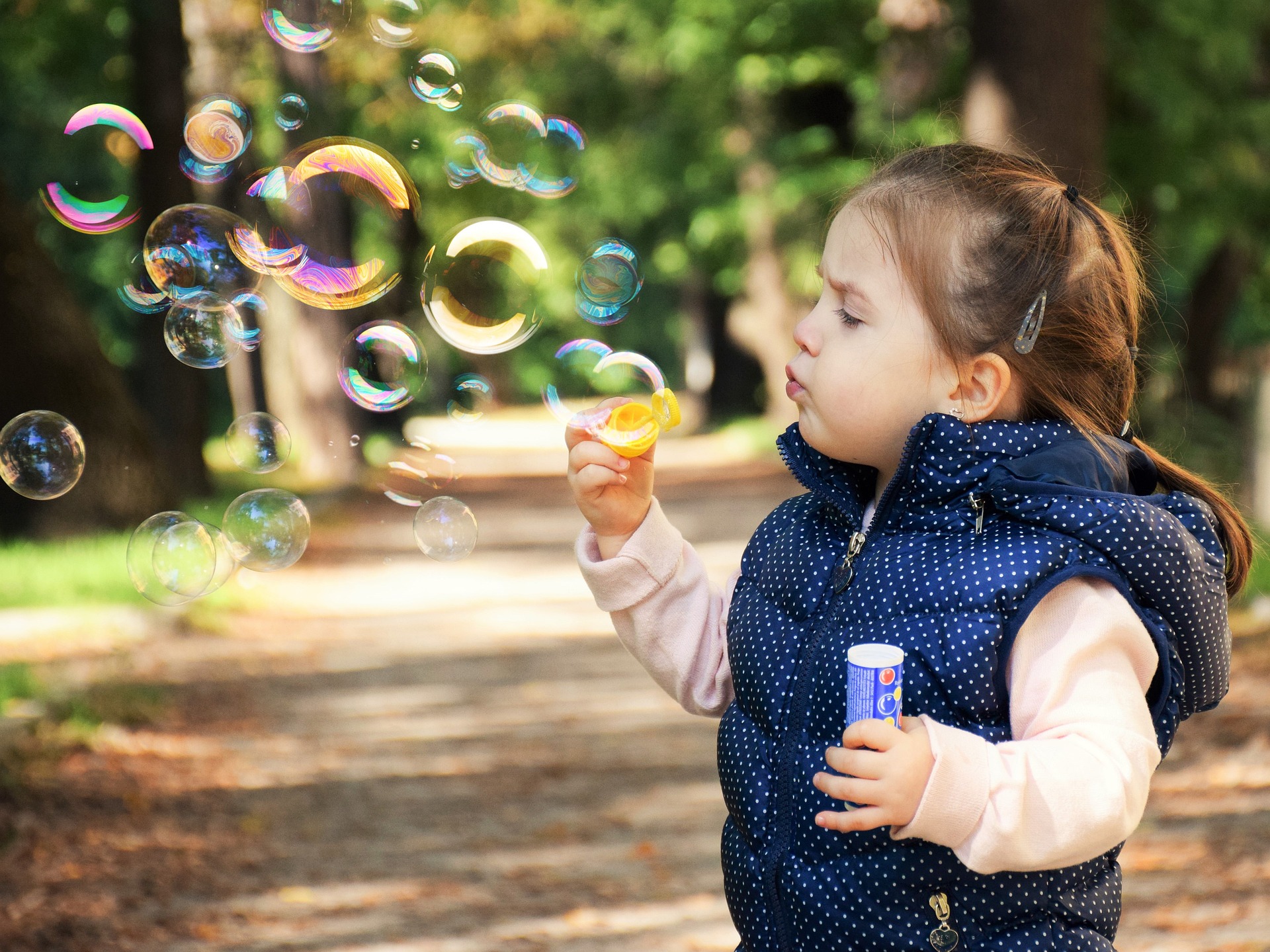 A small girl in a blue spotty waistcoat earnestly blow bubbles from a pink bubble wand. In her other hand, she holds tight to a bottle ob bubble mixture. She is standing on a woodland path and has made a huge mass of bubbles which are catching the light and are edged with rainbow colours.