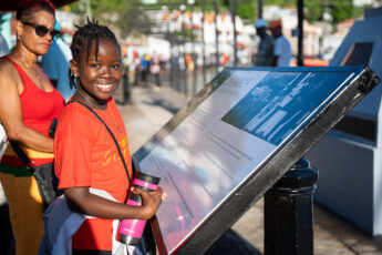 A young girl with her hair in tightly tied braids smiles at the camera. She stands beside an interpretative board on the quad in a port in Grenada. She is wearing a bight orange T shirt and holding a bright pink water bottle. Behind her the blurred images of crowds throng the harbour wall which is edged by the straight trunks of palm trees.