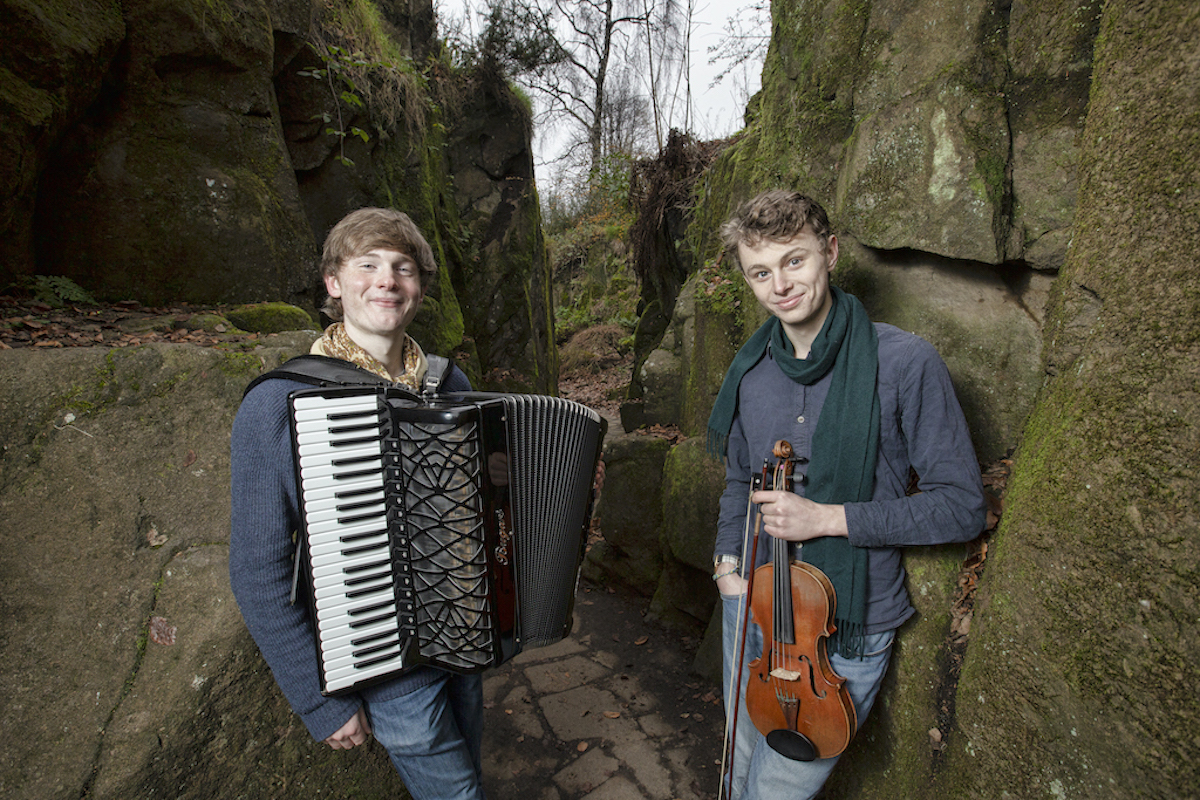 Two cheerfully smiling young men stand among trees leaning against some venerable tree trunks. On the left, he has a piano accordion and on the right a violin. They are dressed casually in jeans and shirts.