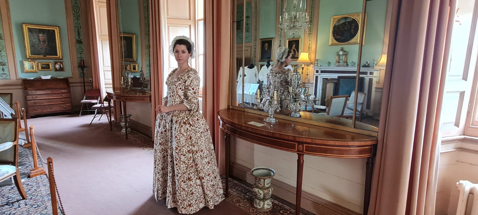 A girl in a mob cap is wearing an Indian print gown in the Georgian fashion standing in an ornate Adam interior