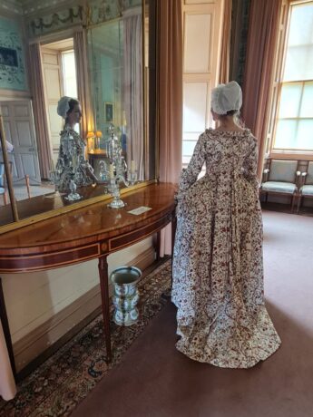 A girl in a mob cap and an Indian print Georgian gown stands with her back to the camera. The dress has full pleats in the back and her reflection is caught in a mirror to her right. The interior is period with a mahogany side table, pair glass and settee.