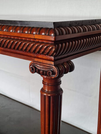 The corner of an ornately carved rosewood table. Beneath a black slate top is a row of carved egg and dart atop two straight bands separated from two similar bands by a deep line of gadrooning. The table leg is columnar, fluted and topped with an ionic capital. The wood is a rich glowing red colour.