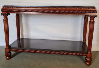 An ornately carved rosewood table with a rich glowing red colour. Beneath a black slate top is a row of carved egg and dart atop two straight bands separated from two similar bands by a deep line of gadrooning. The table leg is columnar, fluted and topped with an ionic capital with a gadrooned ballon at the foot. There is a black slate shelf just above the balloon which echoes the top, both are polished to a sheen.