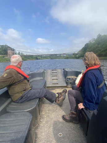 A man and woman in middle age sit at the prow of a boat looking up the river. A suspension bridge crosses the river ahead of them above blue water and banks thick with undergrowth and trees. 