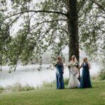 Three girls walk along the river bank on a misty morning. The trees behind them are in full leaf and the water is still and reflective. The girl in the centre is a bride in a white gown and her companions are her bridesmaids dressed in long dark blue stain gowns. All three carry bouquets of flowers and they are laughing together.