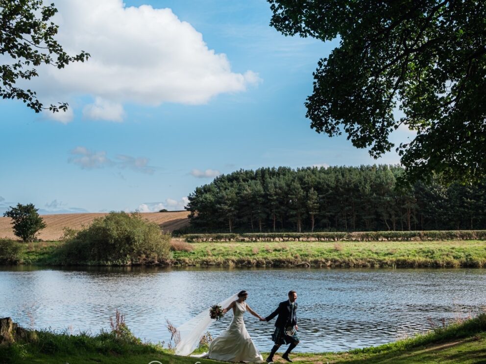 A bride and groom run hand in hand along a woodland path silhouetted against the blue waters of a wide river. On the far bank, a sunlit grassy slope leads into a dark woodland all set off by a blue sky with fluffy white clouds.