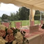 A long table in a marquee is set with a pink cloth and a huge vase of white and pink roses. The chairs are draped with pink and a notice announces the bride and groom/ The side of the marquee is open showing lawns and woodland beyond.