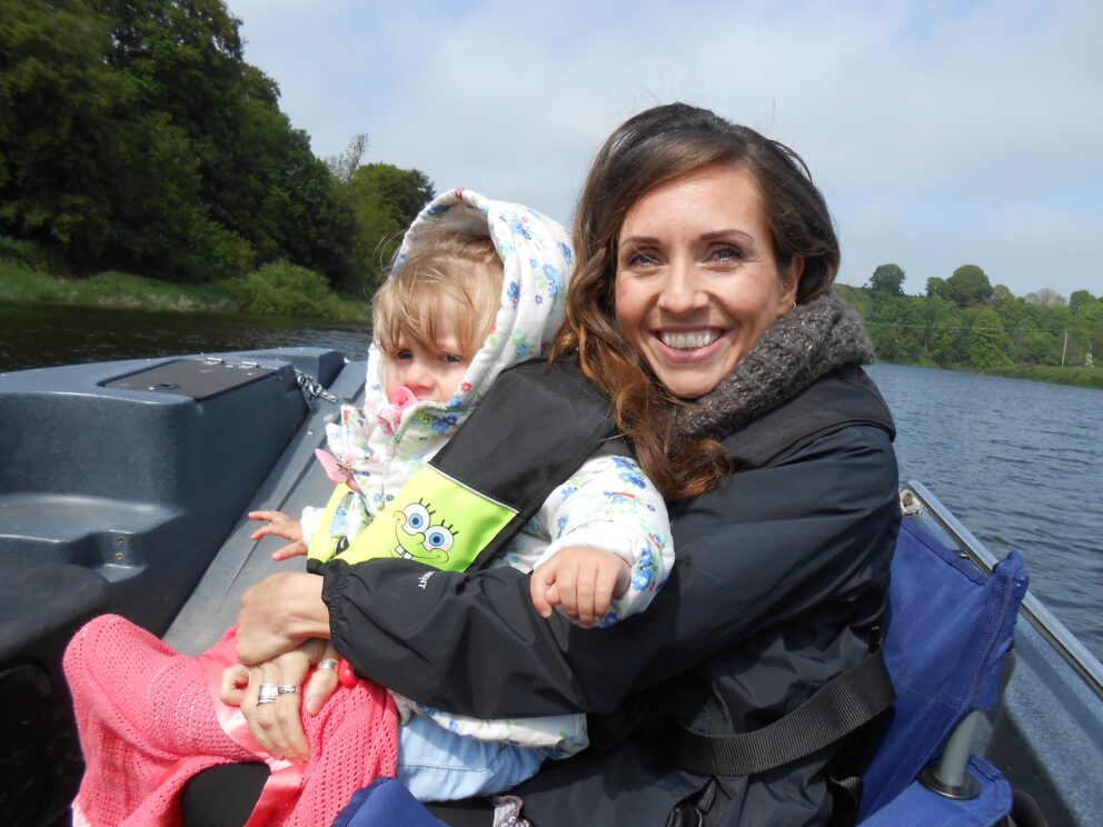 A smiling dark haired girl with a baby on her lap is sitting in a small boat. Behind her is the blue water of the river and the banks on each side are tree lined. She is wearing a dark anorak and the baby, who is around two, is fair haired and wearing pink trousers and a light coloured hooded anorak.