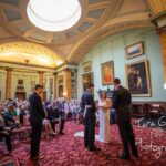 A bride and uniformed groom in the foreground talk to the minister lighted by daylight falling from a glass cupola in the ceiling, The pink patterned carpet leads the eye back to neat rows of guests watching the proceedings. The room is classically decorated with oil portraits hung on green walls divided by yellow scagliola columns.