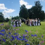 A wedding service is laid out on the lawn behind a bed of brilliant blue flowers. The bride and groom in traditional wedding attire stand in the back ground surrounded by their guests sitting in two blocks on white chairs. The lawns are brilliantly green and the sky brilliantly blue.