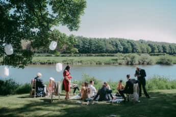 A group of a dozen smartly dressed peoples or sat on rugs around the edge  of the river. The water is blue, the sky clear and the trees on both baks green and verdant. In the trees behind the guests hand white Chinese lanterns suggesting a wedding.