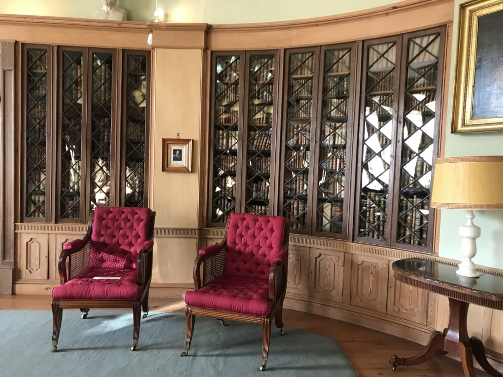 An interior with glass fronted bookshelves on a curving wall. In front are two red upholstered regency armchairs with mahogany arms and legs. The sis the Library at Paxton House