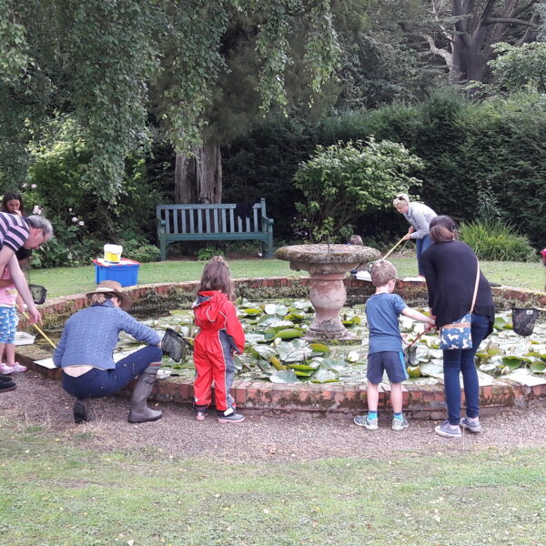 A group of adults and children pond dipping at Paxton House. The pond is circular with a small wall surrounding. There is a fountain in the middle of the pond.